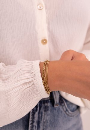 Bracciale in oro con una combinazione di design a catena e accenti delicati, indossato su un polso con una blusa bianca texture.