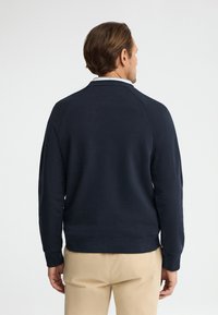 Hombre de pie de espaldas, vistiendo una sudadera azul marino de manga larga y pantalones beige frente a un fondo claro y liso.