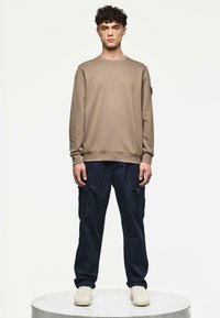 Lichtbruine sweatshirt met geribbelde manchetten en onderrand, gecombineerd met marineblauwe cargopantalons met zijzakken. Model staat op een lichtgrijze voetstuk.