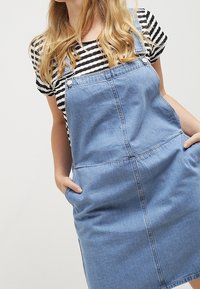 Džínová šaty overall v světle modré barvě, s nastavitelnými ramínky, předními kapsami a detaily šití. Nosené přes černé a bílé pruhované tričko.