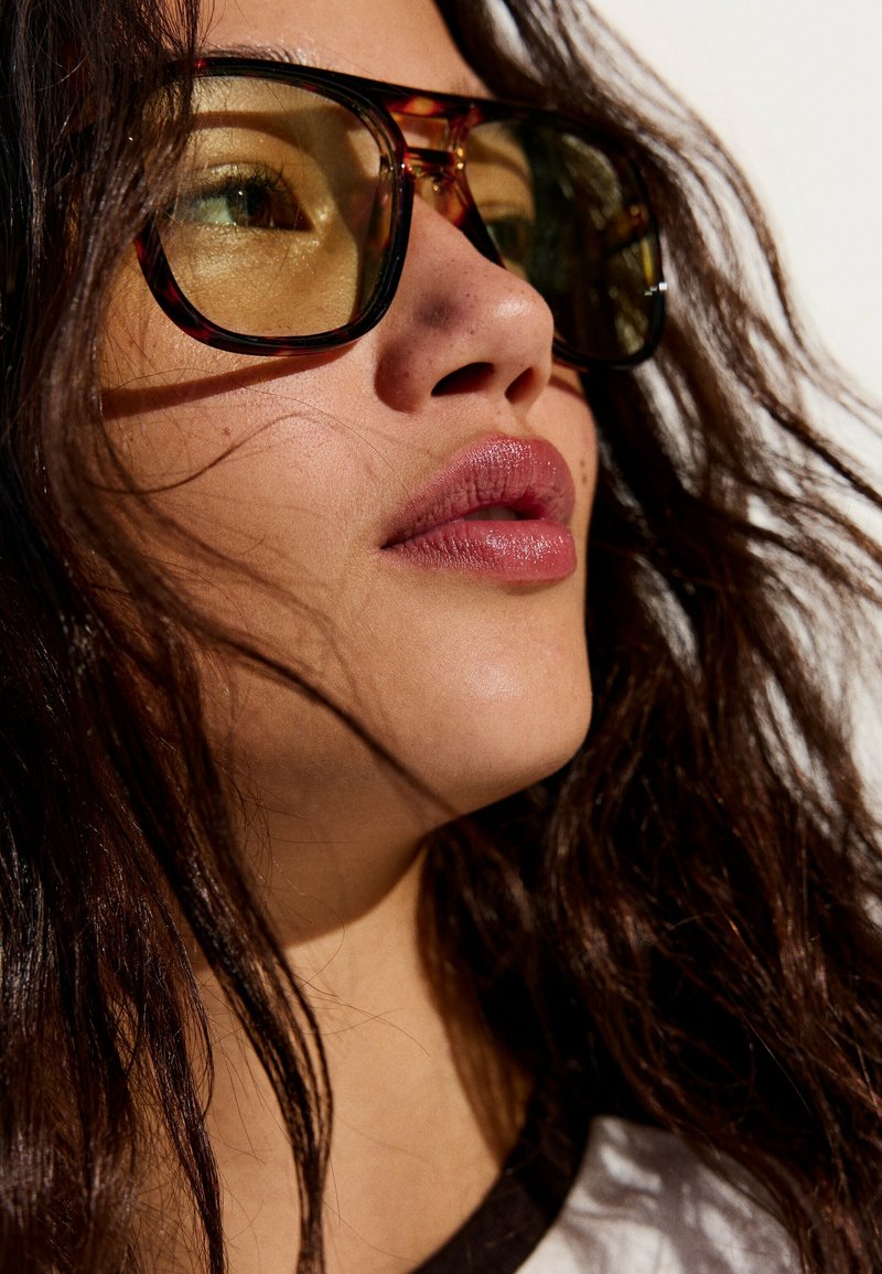 Gros plan d'une personne aux longs cheveux foncés portant de grandes lunettes teintées marron, regardant légèrement vers le haut avec une expression neutre.