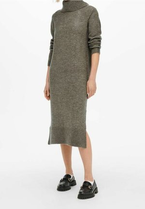 Dzianinowa sukienka swetrowa w kolorze oliwkowej zieleni o długości do kolan, z golfem i bocznymi rozcięciami, zestawiona z czarnymi mokasynami i białymi skarpetkami.