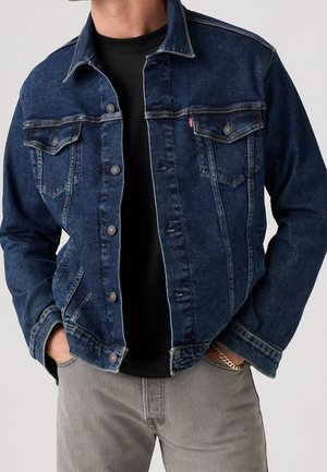 Homme portant une veste en jean bleu foncé sur une chemise noire, les mains dans les poches, associé à un pantalon gris clair et un bracelet chaîne en or.