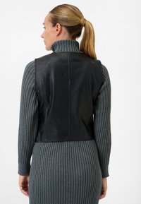 Schwarze, ärmellose Lederweste mit hohem Kragen, getragen über einem taillierten grauen Rippkleid, das die glatte Textur und das taillierte Design betont.