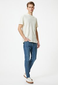 T-shirt a maniche corte beige chiaro abbinato a jeans blu e sneaker bianche con suola marrone. Design semplice, vestibilità rilassata, accenti minimi.