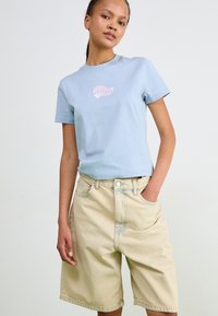 Ljust blå bomullströja med en rosa grafik, kombinerad med beige denimshorts med avslappnad passform och synliga sömdetaljer.