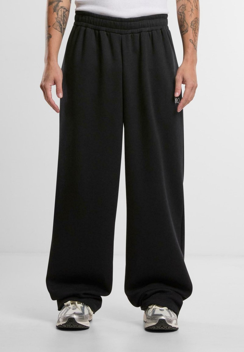 Melni sporta bikses ar elastīgu vidukli, platām kājām un nelielu logo priekšpusē. Izgatavotas no mīksta, teksturēta auduma. Kombinētas ar pelēkiem sporta apaviem.