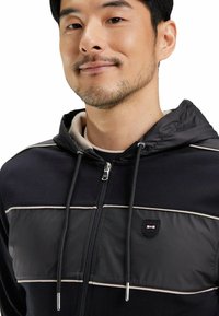 Hoodie zippé noir et marine avec un panneau matelassé, des rayures blanches contrastantes, une capuche avec cordelette et un patch logo sur la poitrine.