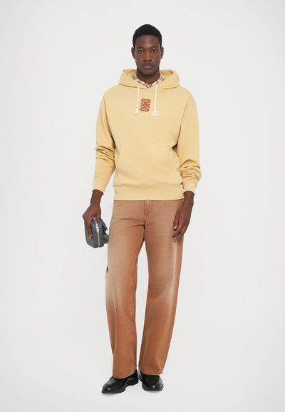 Mann iført en beige hettegenser og brune vide jeans, som holder en liten grå bag, står mot en enkel hvit bakgrunn.