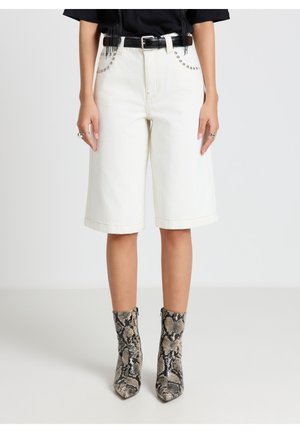 Person trägt weiße Shorts bis zum Knie mit genähten Taschen, schwarzen Gürtel, schwarzen Oberteil und knöchelhohe Stiefel mit Schlangenprint vor einfarbigem Hintergrund.