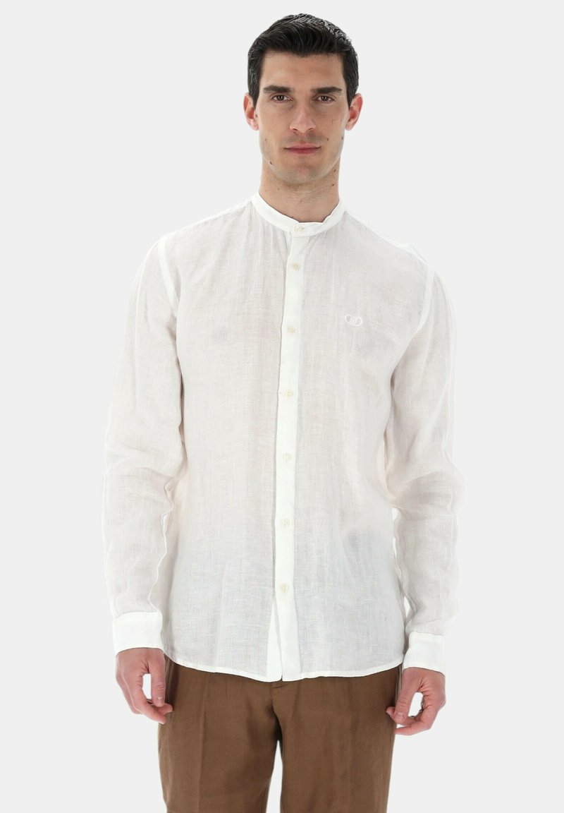Homme portant une chemise blanche à manches longues boutonnée avec col mao et un pantalon marron, debout devant un fond clair uni.