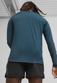 Mėlynas ilgomis rankovėmis sportinis marškinėliai su apvaliu hemu, plokščiomis siūlėmis ir užtrauktuku kišenėje, derinami su juodais šortais, kuriuose yra logotipas.