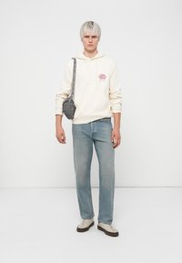 Light beige hoodie with red graphic, light blue straight-leg jeans, gray crossbody bag, and cream sneakers with brown accents.