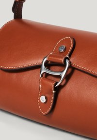 Ruda odinė rankinė, turinti kreivą formą, sidabriniai priedai ir balti siuvinėjimo elementai. Uždarymas turi elegantišką metalinį užsegimą.