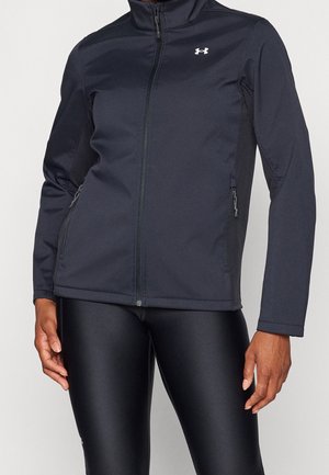 Person, die eine schwarze Under Armour Jacke mit Reißverschluss und schwarze Leggings trägt, steht mit locker an den Seiten hängenden Händen.