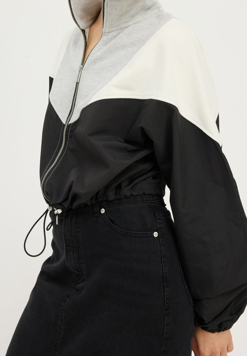 Femme portant une veste zippée color-block en gris, blanc et noir avec une taille élastique, et une jupe en denim noire taille haute.