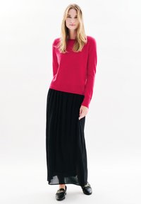 Maglione a maglia fucsia con maniche lunghe, abbinato a una gonna maxi plissettata nera. Modella che indossa mocassini neri con dettagli in catena d'oro.