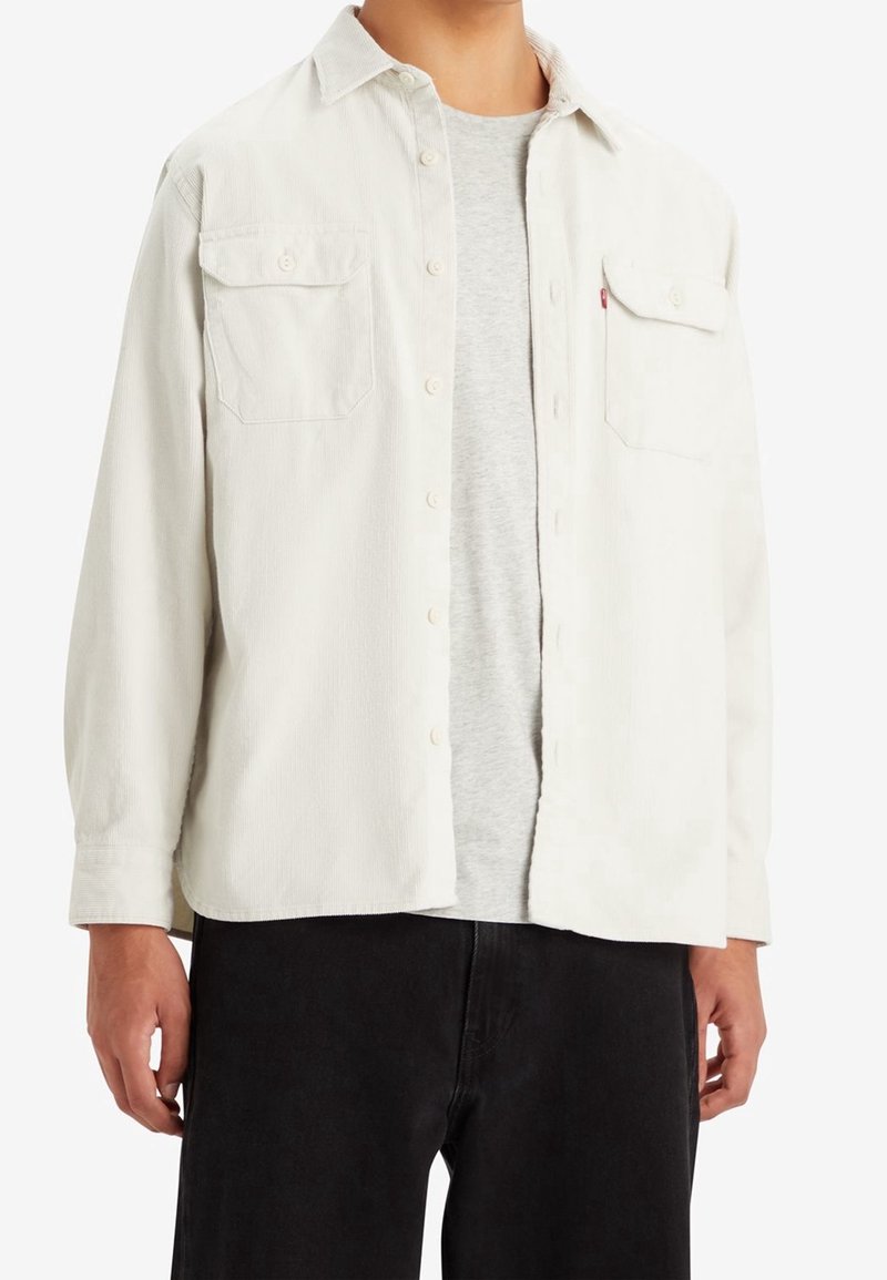 Homme portant une chemise boutonnée ouverte blanc cassé avec des poches poitrine par-dessus un t-shirt gris clair et un pantalon noir.