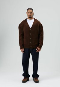 Cardigan marron tricoté avec un col en V, cinq boutons et des poches plaquées avant, associé à un jean bleu à coupe ample et des bottines marron à lacets.