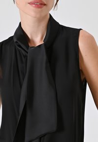 Zwarte mouwloze blouse met een gestrikte kraag. Glad, lichtgewicht materiaal met een minimalistisch ontwerp, met een subtiele drape en zonder zichtbare sluitingen.