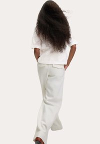 Witte textuur blouse met bloemmotief, gecombineerd met een wijde crèmekleurige broek. Het model heeft lang krullend haar en is van achteren gezien.
