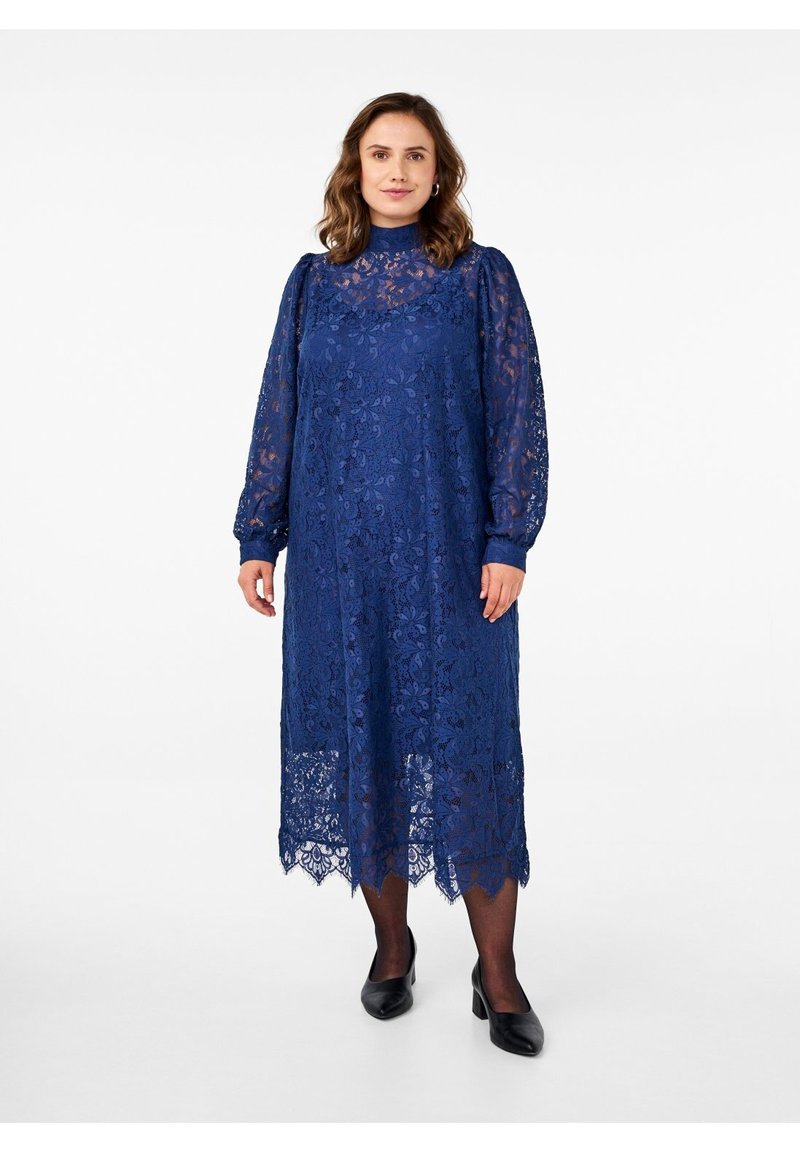 Mujer con un vestido de encaje azul marino de manga larga, cuello alto y largo hasta la rodilla, tacones negros y medias transparentes, de pie frente a un fondo liso.