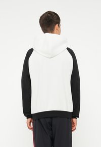 Sweatshirt com capuz apresentando um corpo branco e mangas raglan pretas. Tecido suave com um ajuste relaxado e capuz com cordão, sem logotipo visível.