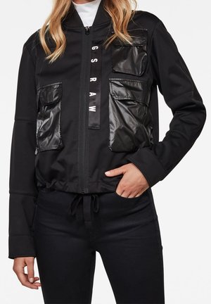 Kobieta ubrana w czarną kurtkę zapinaną na zamek z błyszczącymi kieszeniami oraz pionowym napisem „G S R A W” z przodu, w połączeniu z czarnymi spodniami.