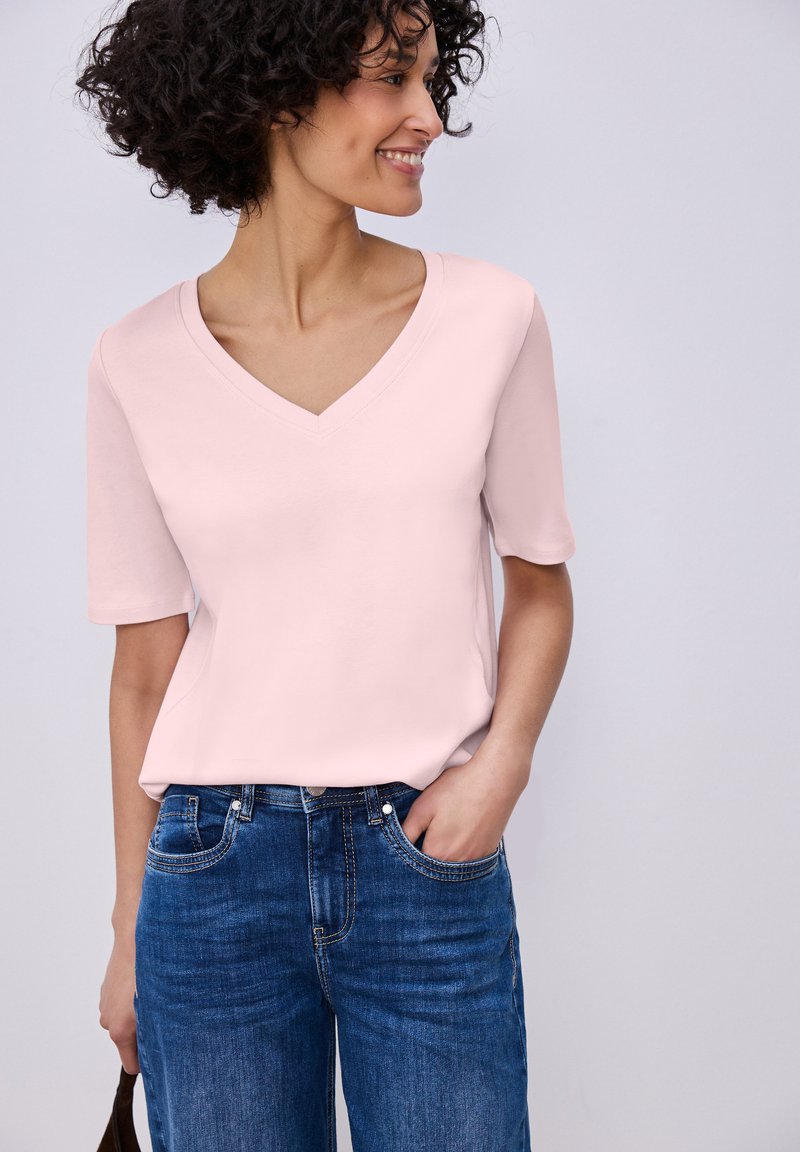 Femme aux cheveux bouclés portant un haut rose clair à manches courtes et col en V, et un jean bleu, souriant et regardant de côté avec une main dans la poche.