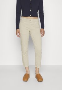 Pantalones recortados beige con un ajuste elegante, bolsillos frontales y cierre de botón, combinados con una blusa de manga larga azul marino y zuecos marrones.