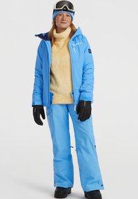 Blauwe geïsoleerde skijas met een capuchon en ritszakken, gecombineerd met blauwe broeken. De outfit heeft een fuzzy beige binnenlaag en zwarte handschoenen.