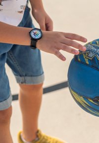 La main d'un enfant tendant la main vers un ballon de basket bleu et jaune tout en portant une montre en cuir noire avec un cadran coloré.