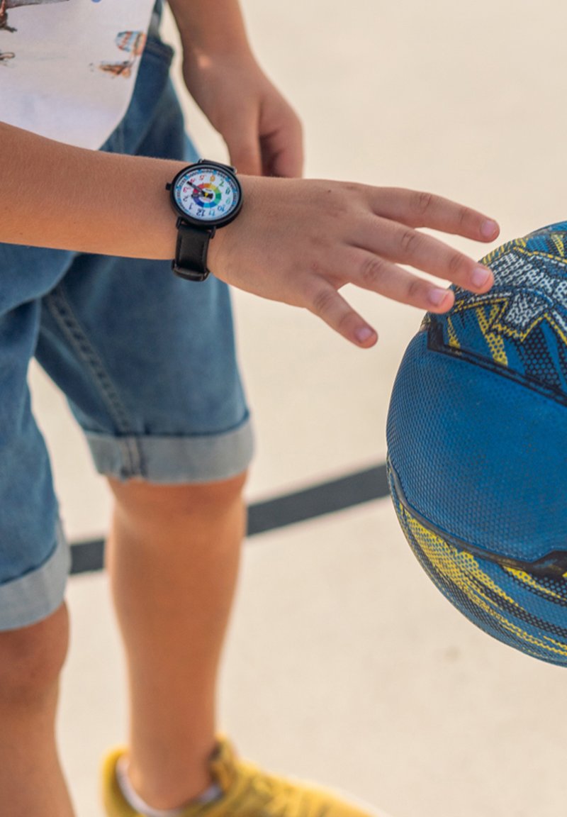 La main d'un enfant tendant la main vers un ballon de basket bleu et jaune tout en portant une montre en cuir noire avec un cadran coloré.