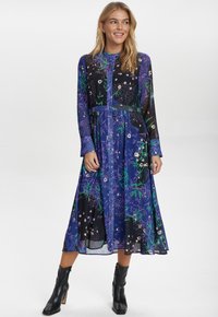 Langdurige, halflange jurk met een combinatie van paarse en zwarte stoffen, bloemmotieven en een aansluitende tailleband. Gecombineerd met zwarte enkellaarsjes.
