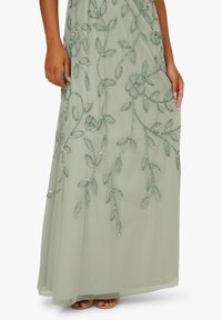 Długa zielona sukienka z kwiatowym haftem i koralikowymi akcentami, charakteryzująca się zwiewnym krojem i delikatnymi wzorami liści na materiale.