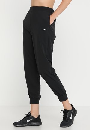 Osoba nosząca czarne spodnie dresowe Nike z elastycznymi ściągaczami i czarne sneakersy Nike, stojąca z jedną ręką w kieszeni na białym tle.