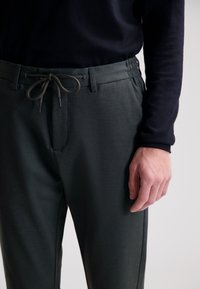 Mørkegrønne, teksturerede bukser med elastisk talje, snøre-lukning og sidelommer. Båret med en mørkeblå bluse med lange ærmer.