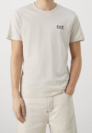 Hombre vestido con una camiseta EA7 Emporio Armani de color beige claro y pantalones a juego con costuras negras, de pie con una mano en el bolsillo.