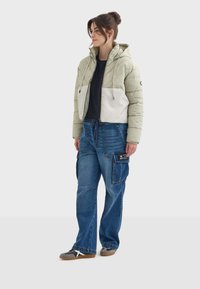 Chaqueta de plumas verde claro con un panel blanco, combinada con jeans cargo azules que tienen bolsillos y una etiqueta negra. Zapatillas grises completan el look.