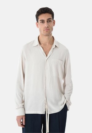 Giovane uomo che indossa una camicia bianca larga con taschino sul petto e pantaloni blu navy, in piedi davanti a uno sfondo bianco neutro.