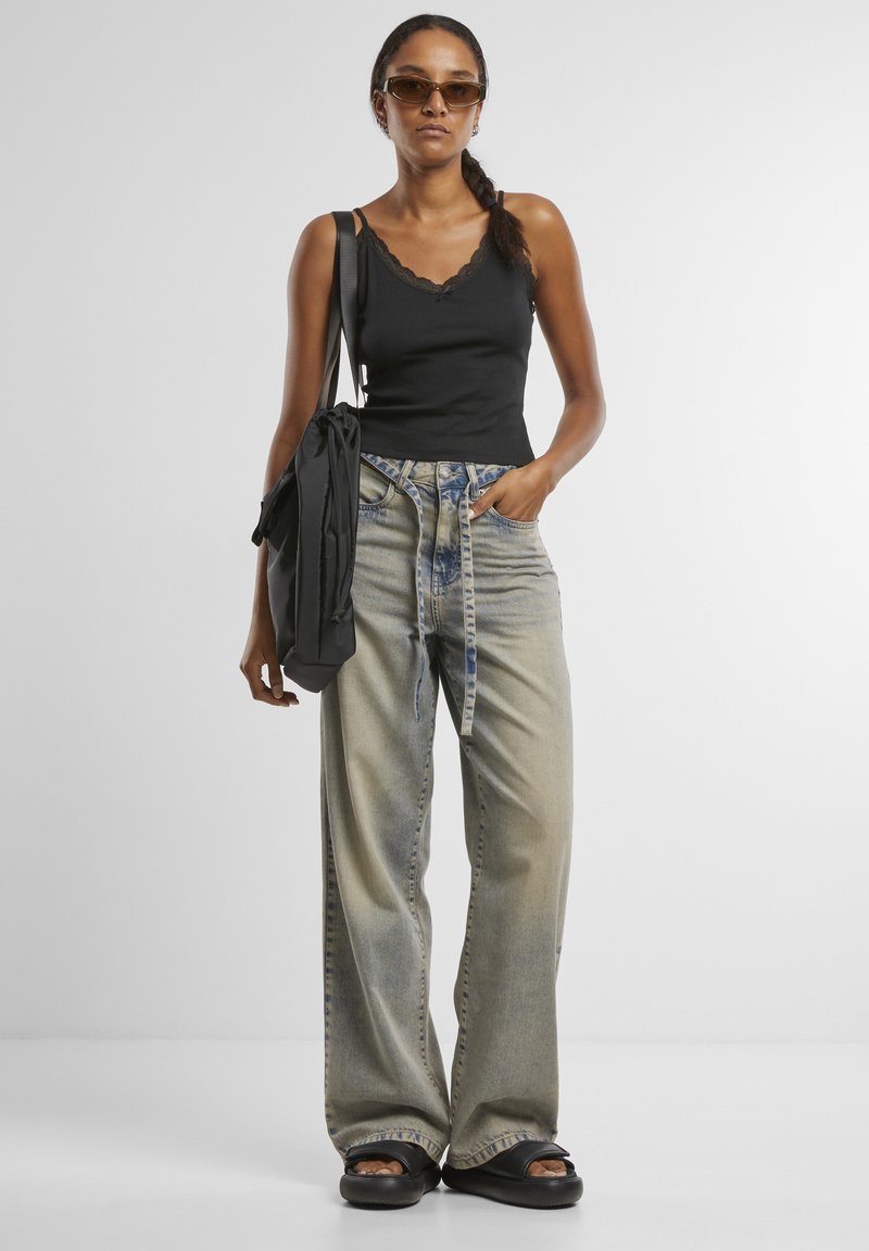 Femme portant des lunettes de soleil foncées, un top noir sans manches, un jean large et délavé avec ceinture, des sandales noires et un sac à épaule noir.