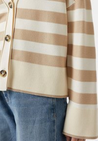 Pasiasty kardigan w kolorach kremowym i beżowym, z dużymi guzikami, prążkowanym wykończeniem na dole oraz rozkloszowanymi rękawami. Materiał wydaje się miękki i dzianinowy.
