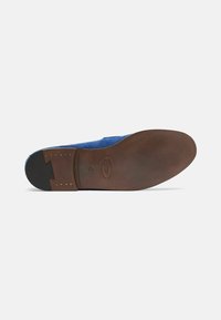 Wildlederschuh in lebhaftem Blau mit einer braunen Ledersohle. Bemerkenswerte Details sind eine Einkerbung in der Ferse und kleine goldene Nieten entlang der Sohlenkante.