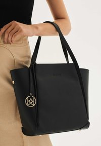 Juoda odinė didelė rankinė su dviem ilgais rankenėliais, puošta auksine pakabuka ir įspaustu logotipu. Fone matoma šviesiai smėlinio spalvos tekstūruota medžiaga.