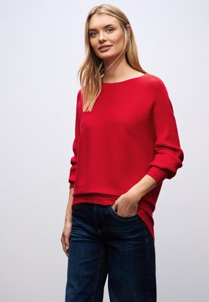 Roter gerippter Pullover mit weitem Ausschnitt und langen Ärmeln, kombiniert mit dunkelblauen Jeans. Der Pullover hat eine lockere Passform und eine strukturierte Oberfläche.
