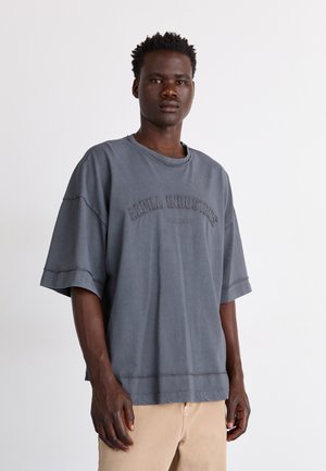 Młody mężczyzna w luźnej, wyblakłej ciemnoszarej koszulce z napisem „Alpha Industries” oraz beżowych spodniach, stojący na jednolitym tle.