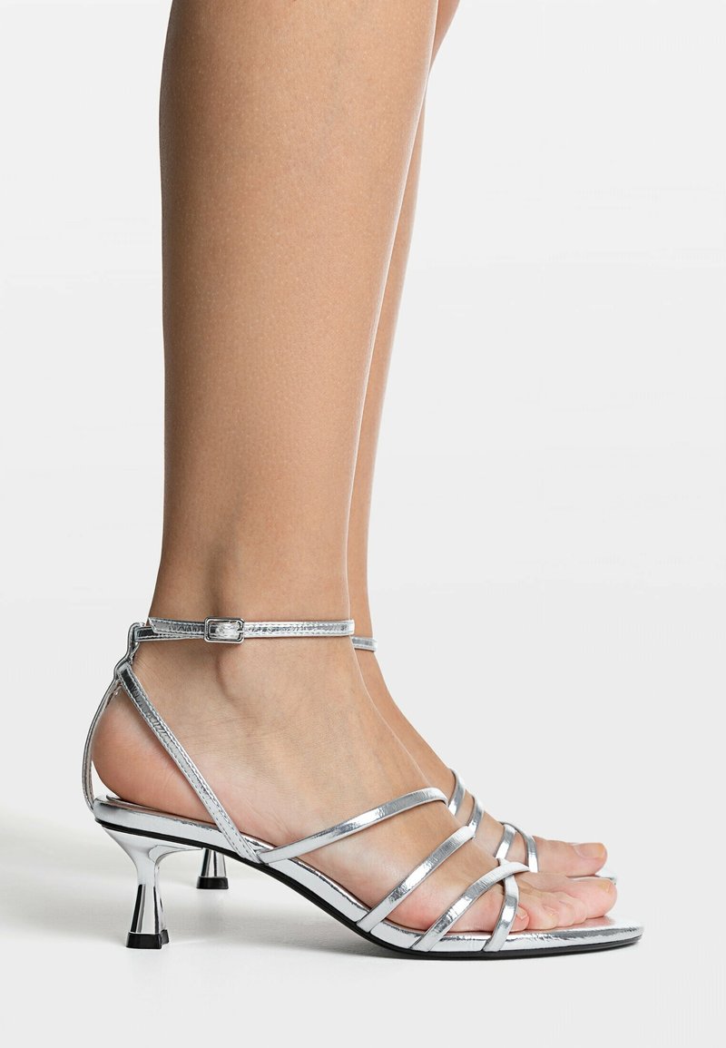 Sandales à talons kitten argentées brillantes avec brides et boucles à la cheville, portées pieds nus sur un fond uni.