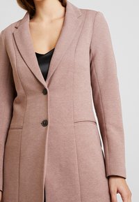 Jasnoróżowy, dopasowany płaszcz o usztywnionej konstrukcji, z pojedynczym zapięciem na guzik, klapami z wcięciem i bocznymi kieszeniami; widoczna faktura tkaniny.