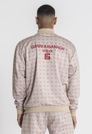 Chaqueta bomber de color beige claro con patrón de logotipos repetido, puños y dobladillo acanalados, que presenta el texto rojo "Gianni Kavanagh Atelier" en la parte posterior.