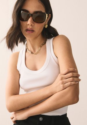 Mujer con grandes gafas de sol con montura carey, collar de cadena dorada y pendientes, camiseta sin mangas blanca y pantalones negros, posando con los brazos cruzados.